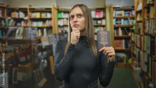 Photos Young woman holding czech passport with hand on chin in bookstore pondering travel decisions; pensive doubt
