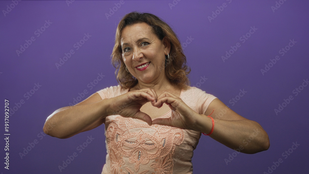 Obraz premium Hispanic middle aged woman with soft smile making heart shape gesture with hands in purple studio; love.