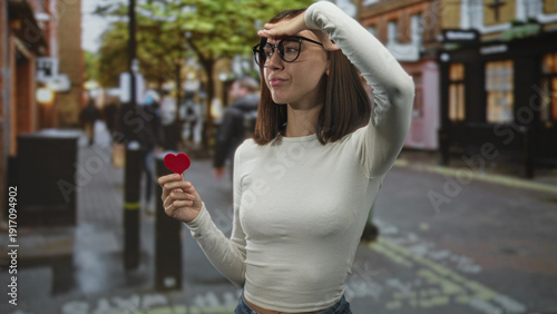 Φωτογραφία Young woman wearing glasses and a white long sleeve top holds a small red heart and shields her eyes with hand on forehead on a street lined with shops; longing romance
