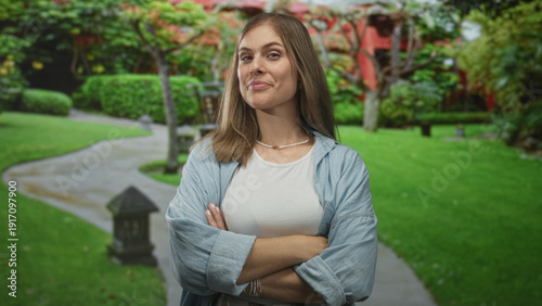Εκτύπωση καμβά Woman standing with arms crossed and smirking on lush green grass in front of red building with trimmed hedges; rejection