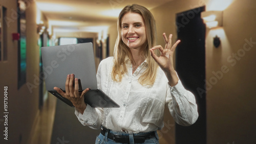 Quadro su tela Blonde woman holds laptop, smiling and making ok sign with hand while standing in a hotel building corridor, wearing white blouse and jeans; confidence success