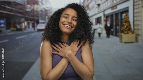 Young hispanic woman extends arms toward camera in street with a welcoming gesture; friendliness warmth.