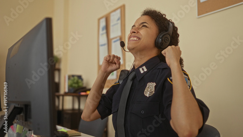 Woman celebrating in police uniform at station office, appearing excited with fists raised, seated at desk with computer, wearing headset in professional indoor workplace setting.
