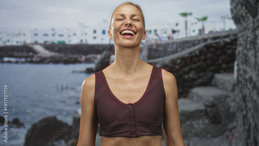 Fototapeta premium Woman smiles and tilts head, showing exposed midriff and maroon tank top in front of a seaside building with volcanic rocks; joy.