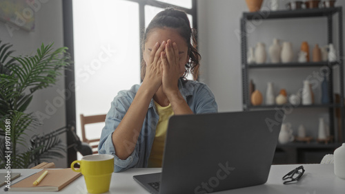 Obraz na płótnie Woman covers face with hands at laptop in studio, seated at a cluttered desk with yellow mug, notebook, glasses and potted plant; stress fatigue