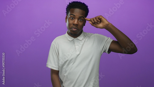 Fotografi Young african american man in white polo shirt points finger to temple in studio against purple studio wall; playful madness