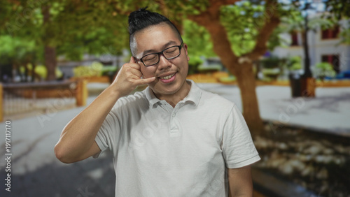 Man with hand to ear in call gesture on street wearing white polo and glasses; friendly greeting connection.