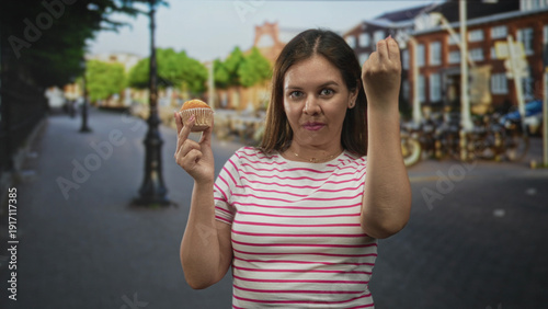 Woman in striped t shirt holds muffin in one hand and makes a fist gesture with the other on a city street; hunger annoyance. Free Download Woman in striped t shirt holds muffin in one hand and makes a fist gesture with the other on a city street; hunger annoyance. - GFXLoad