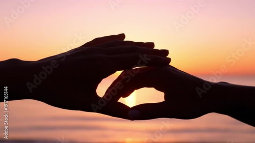 Silhouettes of two hands forming a heart shape against a beautiful sunset over the ocean, symbolizing love and connection.