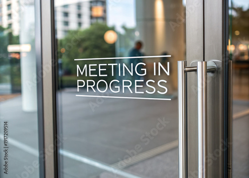 Meeting in progress sign on a glass office door with silver handle and blurred city street background with trees and buildings
