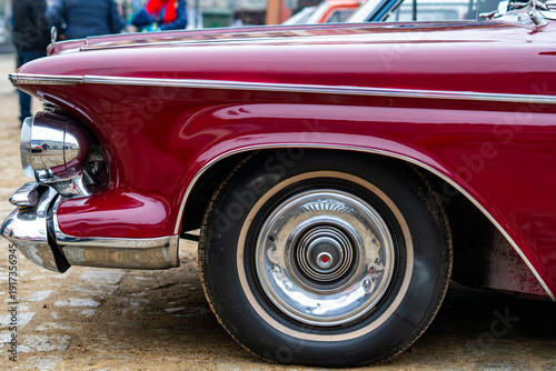 Classic maroon vintage car with chrome bumpers and whitewall tires, close-up of the front fender and stylish hubcap during an antique automobile show in the city.