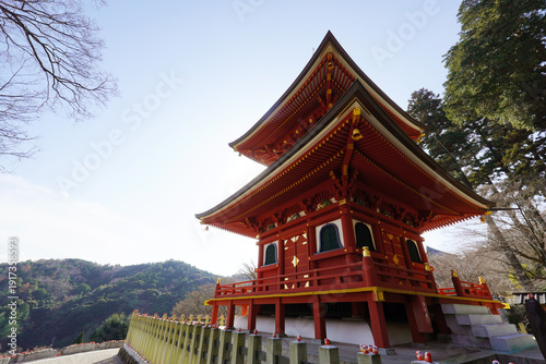 Stunning vibrant red two-story Japanese Buddhist pagoda at Katsuo-ji Temple in Osaka. Traditional temple architecture set against a lush mountain forest backdrop under a clear blue sky. Symbol of peac