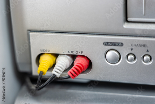 Close up of RCA composite output on VCR front panel. Yellow video and red white audio connectors plugged into analog AV output ports. Retro home electronics detail.