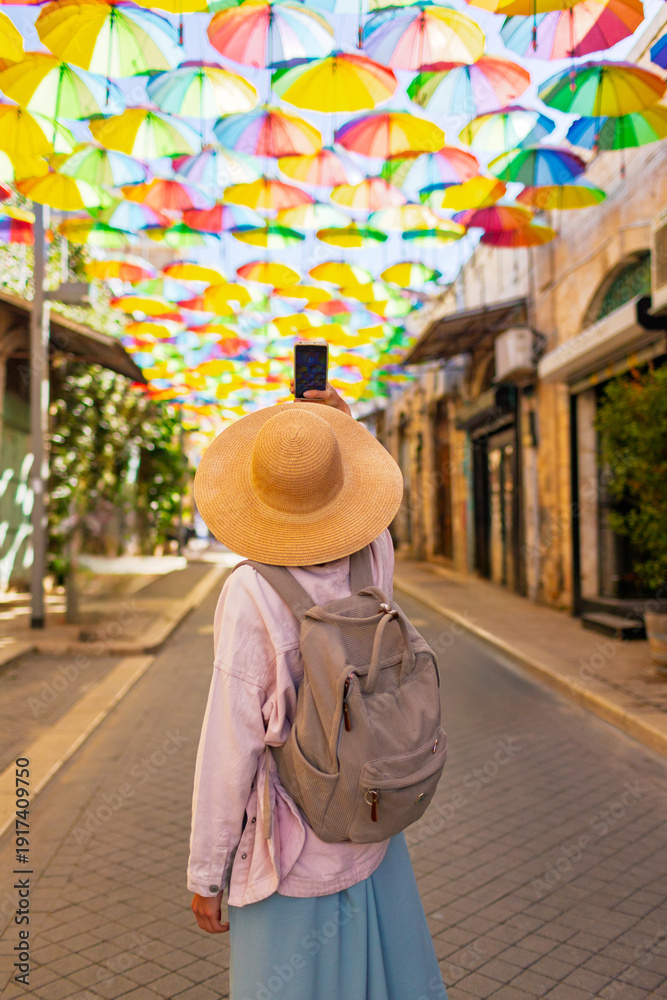 Fototapeta premium Girl Tourist on an excursion stands on the street with hanging umbrellas and takes pictures. umbrella sky.