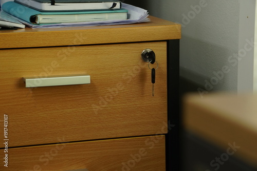 Soft, diffused natural light illuminates a wooden office desk, highlighting the simple details of a locked drawer and a stack of notebooks.