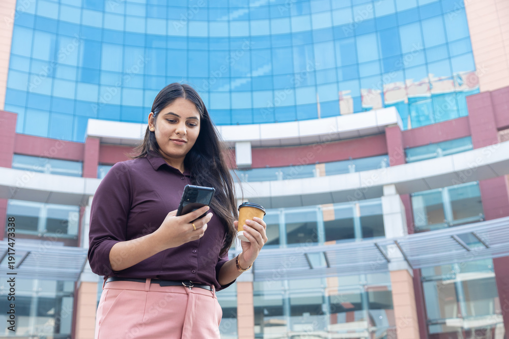 Fototapeta premium Young indian businesswoman checking her smart phone and holding a disposable coffee cup standing outside a modern corporate office building. Corporate job. Copy space. Technology.