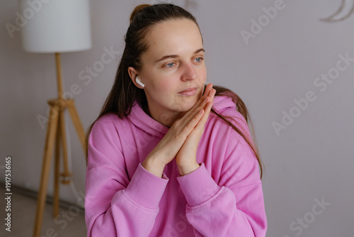 A woman is indoors in a cozy space wearing a pink hoodie. She is listening to music while resting her hands on her chin and looking thoughtful