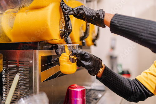 Photos Person in black gloves pours mango smoothie from a slush machine into a clear cu