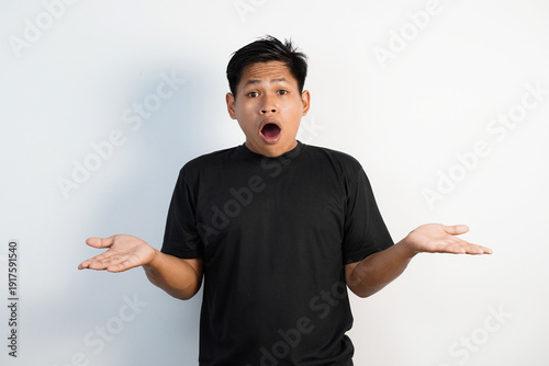 Portrait of a young Asian man in black shirt shrugging with a confused face against a white background, conveying uncertainty and casual demeanor in an expressive body language and candid setting.
