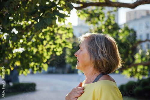 Wallpaper Mural Senior woman with short gray hair wearing a yellow shirt stands outdoors, sunlight filtering through green leaves in a park setting, profile view against a blurred background Torontodigital.ca