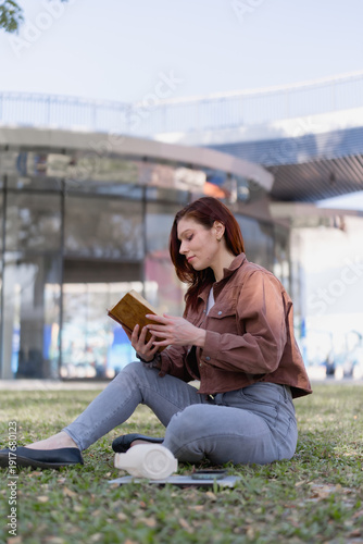 Female university student sitting on a college campus lawn, enjoying a good book outdoors, surrounded by modern architecture, symbolizing education, study, and relaxing downtime