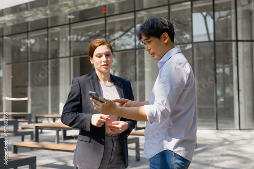 Two business professionals standing outdoors, discussing work and pointing at a smartphone screen, reflecting a concept of modern urban business, communication, and digital technology
