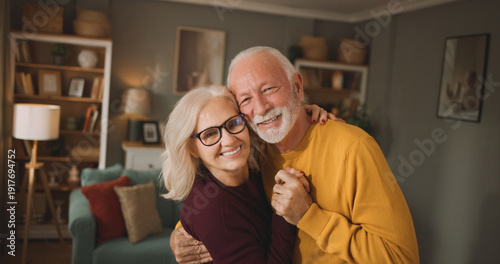 Elderly couple stands close together in their living room. They smile and hold hands while enjoying a warm moment in the afternoon light.