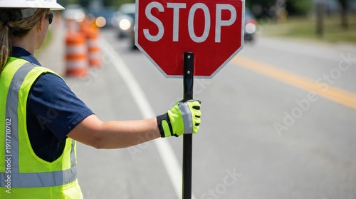 Female traffic controller in safety vest and hard hat holds stop sign while directing vehicles on a road with traffic cones and blurred cars in the background during daytime