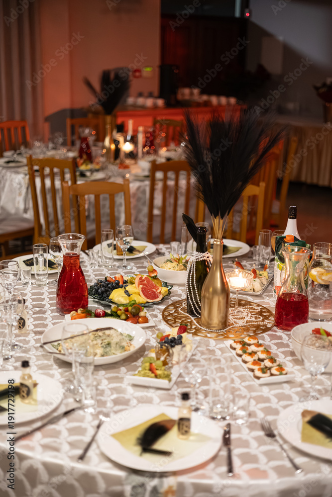 Fototapeta premium Beautifully arranged dinner table with assorted appetizers, fruits, salads, wine bottles, and candles in warm ambient lighting. Decorative centerpiece and elegant tableware create a festive atmosphere