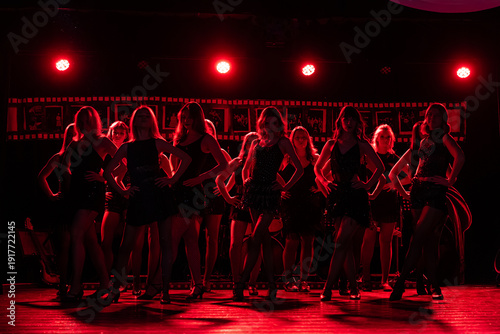 Fotografía Group of female dancers posing confidently on a stage under intense red lighting, wearing black performance outfits and heels