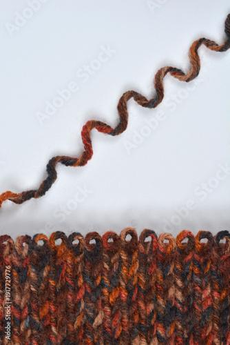 A vertical flat lay shows a dark, multicolored hand-knitted wool swatch with a decorative rolled edge next to a single, wavy strand of matching yarn on a plain gray background