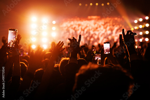 Music festival crowd with raised hands and smartphones under orange lights