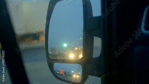 side mirror reflection of headlights at dusk, truck and trailer looming in glass, urban highway scene with bokeh traffic lights, tense evening commute and vigilant driving perspective