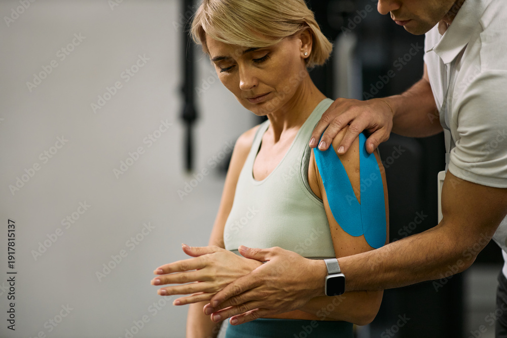 Naklejka premium Middle aged Caucasian woman receiving physical therapy from male therapist applying kinesiology tape to shoulder, focusing on rehabilitation and guided arm movement