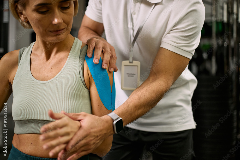 Obraz premium Middle aged Caucasian woman receiving physical therapy from male therapist applying kinesiology tape to shoulder, therapist supporting arm during rehabilitation session in clinic