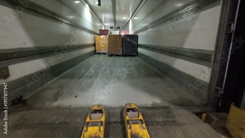 First person point of view shot of an electric pallet jack driving into a semi trailer at a loading dock to pick up a shrink wrapped pallet