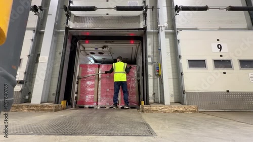 A warehouse worker in a high visibility vest enters the back of a semi trailer at loading dock number nine to remove a load bar before walking away