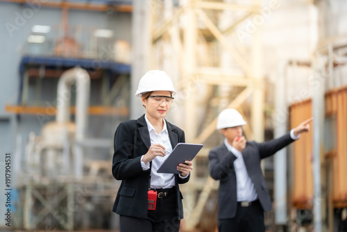 Male and female executives wearing suits and helmets are working within the power plant environment, guiding the industry workforce with a laptop and directing radio communication clearly.