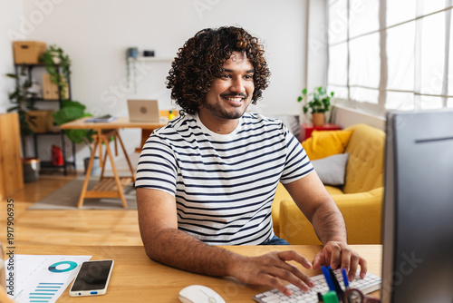 Young Indian man freelancer working on desktop computer at home office desk. Remote professional typing on keyboard and smiling while focusing on online project. Business and working at home concept.