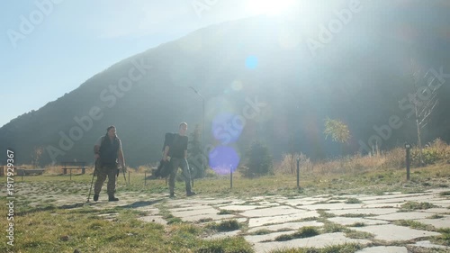 Adult man and teenage boy hikers travel at sunset along rocky mountain path, reaching guesthouse for overnight stay. Two travelers arrive at destination in mountains, ready to rest from fatigue.