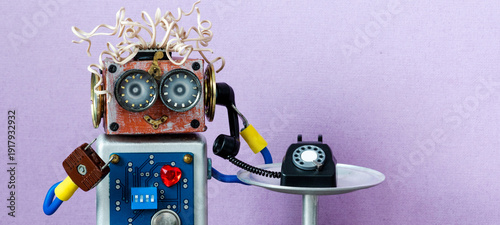 Smiling big-eyed toy robot with wire hair holding vintage rotary phone receiver, listening. Hotline operator concept, customer support advertisement