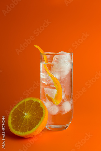An iced cocktail in a Hi Ball Glass with a twist of orange and half an orange against an orange background.