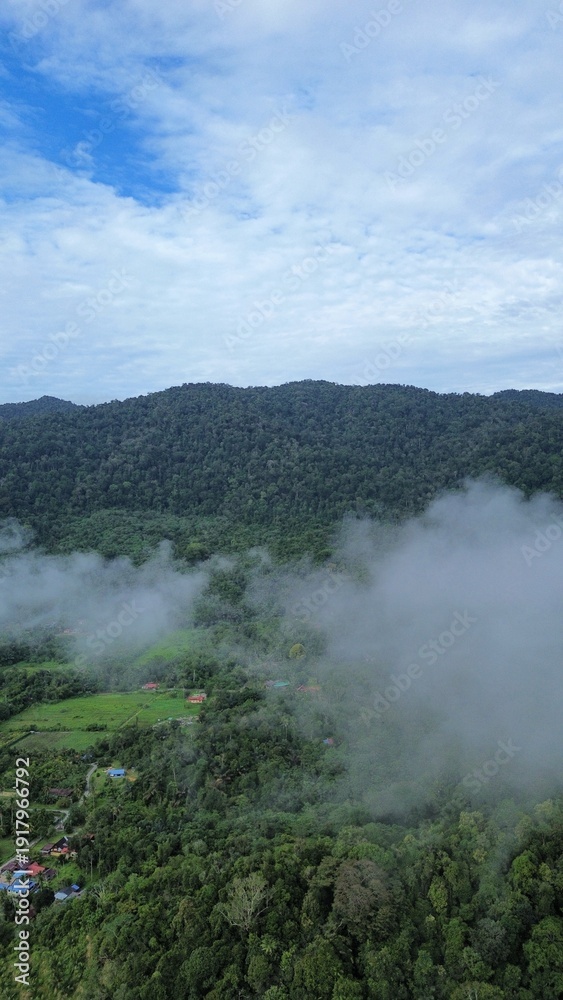 Fototapeta premium Drone aerial view of misty clouds floating over green forest and rural village in a mountainous landscape during early morning.
