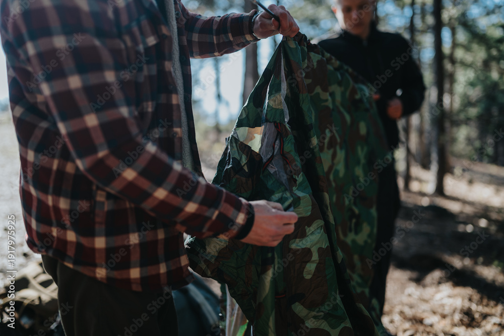Fototapeta premium Two people in outdoor gear unfold and fold a camouflage tent in a wooded area. The scene captures a calm camping moment with natural light and trees in the background.