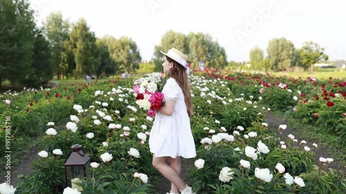 Smiling happy girl holding flowers pink peonies bouquet in a blooming spring garden. Copy space. Beautiful young girl in white dress and straw hat walking with pink flowers in a peony field.  