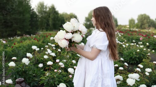Young girl in white dress standing in peony field, holding pink flower bouquet. Smiling and smelling peonies. Summer nature background, romantic lifestyle concept, beauty and femininity. Copy space.