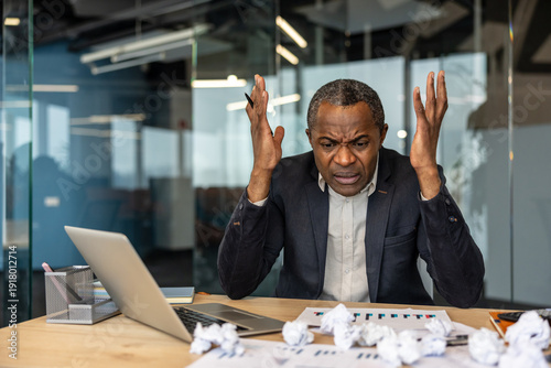 Slika na platnu Mature businessman stressed at his desk, clutching his head over a laptop and cr