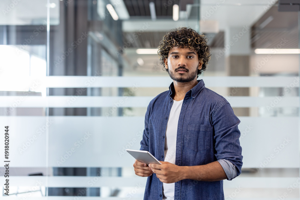 Naklejka premium Portrait of a young Indian man in a shirt standing in an office, holding a tablet and looking seriously at the camera