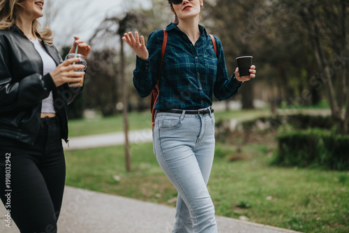 Εκτύπωση καμβά Two women stroll along a park path with coffee cups, casual outfits, and relaxed mood