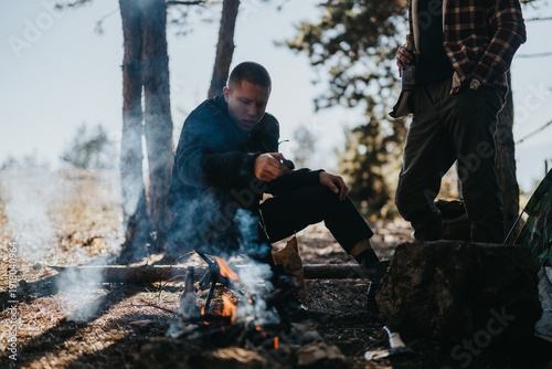 Fotografie Two men gather around a small campfire in a woodland setting; one sits close to the flames